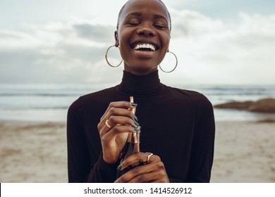 Smiling young african woman standing at the beach with cold drink bottle. Female having soft drink at the sea shore.