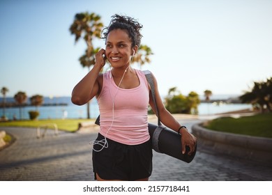 Smiling young African woman in a tank top and shorts listening to music on earphones while walking along a promenade carrying a yoga mat - Powered by Shutterstock