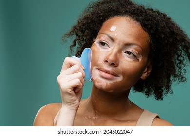 Smiling young african woman with vitiligo condition doing facial massage with gua sha tool on green background - Powered by Shutterstock