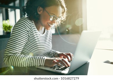Smiling young African woman wearing glasses working online with a laptop while sitting alone at a table