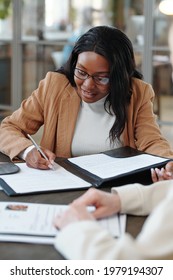 Smiling Young Afro-American Woman Sitting At Table And Signing Contract While Accepting Job Offer After Interview