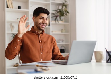 Smiling Young Arab Business Man Making Video Call Using Laptop At Home Office, Having Web Conference Waving Hand At Computer Web Camera And Talking. Online Communication And Remote Work Concept