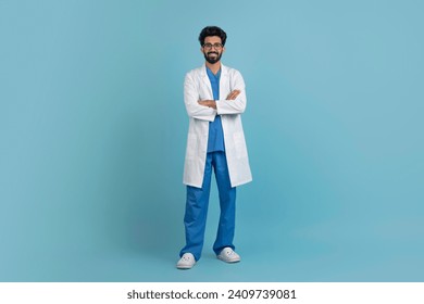 Smiling young arab doctor man posing with folded arms over blue studio background, confident middle eastern male physician wearing uniform and stethoscope, full body length, copy space - Powered by Shutterstock