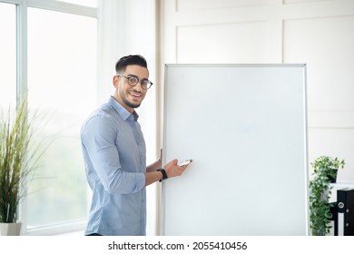 Smiling Young Arab Male Teacher Standing Near Empty Blackboard With Mockup, Giving Online Private Lesson From Home. Millennial Eastern Tutor Conducting Lecture To Remote Students On Web