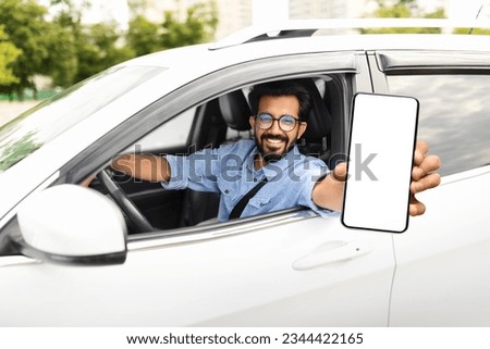 Smiling young arab man sitting in auto, showing smartphone with empty screen, showing newest mobile application for navigation, mockup. Car leasing, automobile, driving and modern technologies