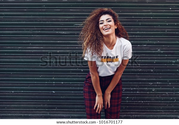 黒い巻き毛の若いアラビア人女性に微笑みかける 通りで普段着のアラブの女の子 白いtシャツを着た幸せな女性と 都市のブラインドにチェックを入れたズボン の写真素材 今すぐ編集
