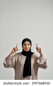Smiling young arabic woman in hijab pointing fingers up while looking upstairs on light background with copy space, vertical shot. Beautiful muslim lady