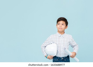 Smiling young asian boy aspiring to be future engineer holding white hardhat and blueprint looking upward in light blue isolated studio backgorund