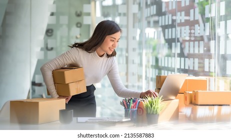 Smiling Young Asian Business Owner Woman Prepare Parcel Box And Standing Check Online Orders Of Product For Deliver To Customer On Laptop Computer. Shopping Online Concept.