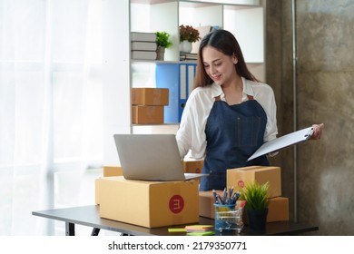 Smiling Young Asian Business Owner Woman Prepare Parcel Box And Standing Check Online Orders Of Product For Deliver To Customer On Laptop Computer. Shopping Online Concept.