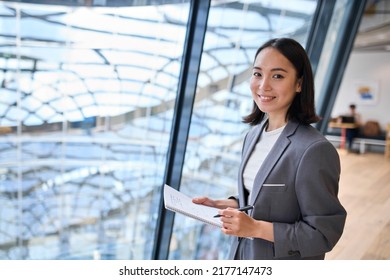 Smiling young Asian business woman manager wearing suit holding notebook standing in modern glass office. Professional executive manager, corporate leader looking at camera. Portrait - Shutterstock ID 2177147473