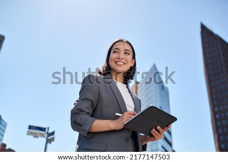 Smiling young Asian business woman leader entrepreneur, professional manager holding digital tablet computer using software applications standing on the street in big city on sky background. Foto stock © 