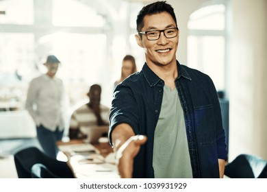 Smiling young Asian businessman extending a handshake while standing in an office with colleagues working in the background - Powered by Shutterstock