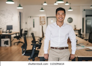 Smiling young Asian businessman leaning against a table while working alone in a large modern office