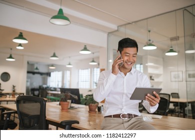 Smiling young Asian businessman sitting on a desk talking on a cellphone and using a digital tablet while working alone in a modern office