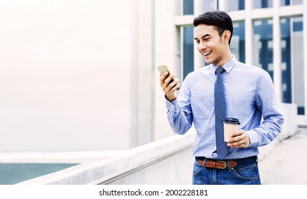 Smiling Young Asian Businessman Using Mobile Phone in the City. Modern Urban Lifestyle. Male Portrait. Hand holding Coffee Cup. looking at Smartphone