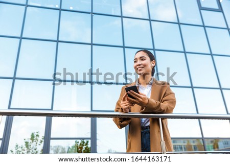 Smiling young asian businessowoman wearing coat standing infront of a glass building outdoors in the city, using mobile phone
