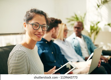 Smiling Young Asian Businesswoman Sitting On A Sofa With A Diverse Group Of Colleagues In A Modern Office