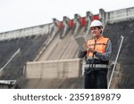 Smiling young Asian civil engineer standing with his arms crossed wearing a helmet and using a walkie-talkie. View and inspect the operation of the dam