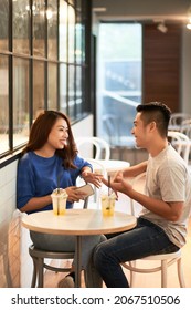 Smiling Young Asian Couple In Casual Outfits Sitting At Small Table With Drinks And Chatting Together In Cafe