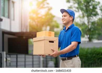 A smiling young asian delivery in blue uniform with parcel cardboard in front of customer house. Messenger and delivery concept.