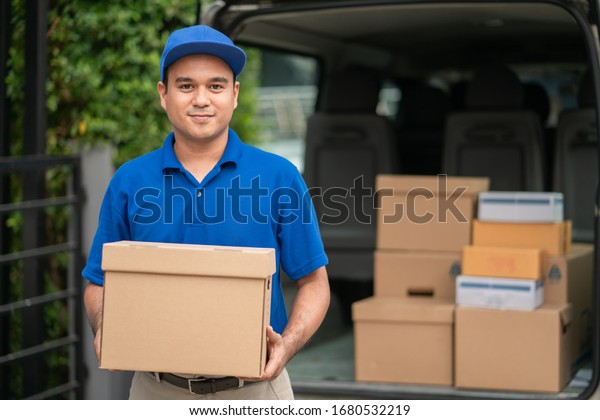 A smiling young asian delivery in blue uniform\
with parcel cardboard in front of customer house. Messenger and\
delivery concept.