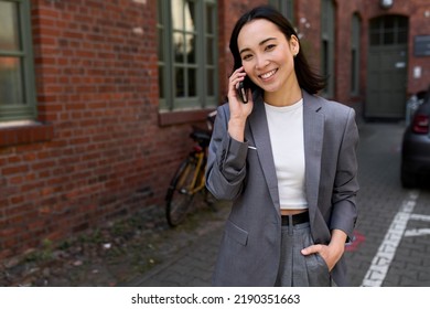 Smiling Young Asian Ethnic Business Woman Professional Manager, Korean Businesswoman Executive Leader Wearing Suit Talking On Cell Phone Making Mobile Business Call Standing Outside Office.