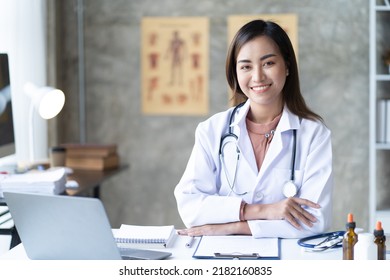 Smiling Young Asian Female Doctor Working At The Clinic Reception, She Is Using A Computer And Writing Medical Reports.