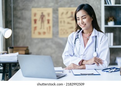 Smiling Young Asian Female Doctor Working At The Clinic Reception, She Is Using A Computer And Writing Medical Reports.