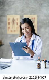 Smiling Young Asian Female Doctor Working At The Clinic Reception, She Is Using A Computer And Writing Medical Reports.