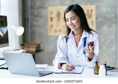 Smiling Young Asian Female Doctor Working At The Clinic Reception, She Is Using A Computer And Writing Medical Reports.