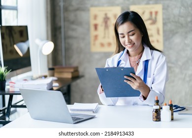 Smiling Young Asian Female Doctor Working At The Clinic Reception, She Is Using A Computer And Writing Medical Reports.