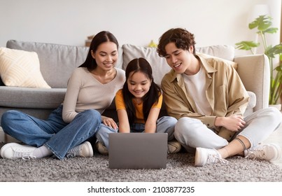 Smiling Young Asian Female, Male And Teen Girl Sitting On Floor And Watching Video, Searching, Browsing On Laptop In Living Room Interior. Video Call Together, Modern Device At Home And New Normal