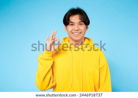 Smiling young Asian guy in a yellow sweatshirt makes a positive 'OK' hand gesture against a blue background