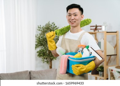 Smiling Young Asian Korean Guy With Bucket Of Detergents With Hands In Rubber Protective Gloves Holding. Cleaning Home Service Concept. Handsome Man Face Camera With Confident Smile Enjoy Housework