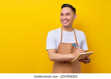 Smiling young Asian man barista barman employee wearing brown apron working in coffee shop write down order menu in notebook look aside isolated on yellow background. Small business startup concept - Powered by Shutterstock