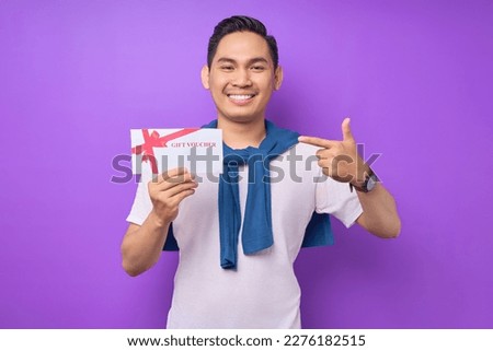 Smiling young Asian man in casual clothes pointing a finger at gift voucher certificate and looking at the camera isolated on purple studio background