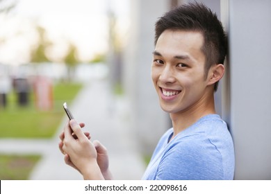 Smiling Young Asian Man Holding Smart Phone And Looking At Camera On City Sidewalk