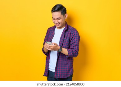 Smiling Young Asian Man In Plaid Shirt Using Mobile Phone Typing SMS Message Isolated On Yellow Background