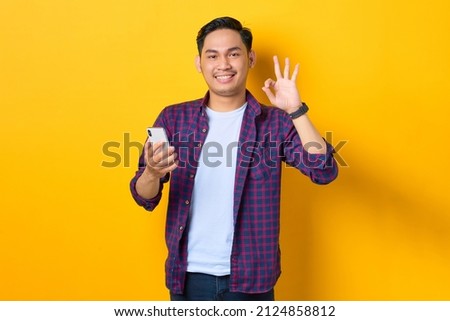 Smiling young Asian man in plaid shirt holding mobile phone and showing okay sign isolated on yellow background