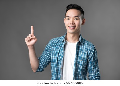 Smiling Young Asian Man Pointing His Index Finger Up To Empty Space