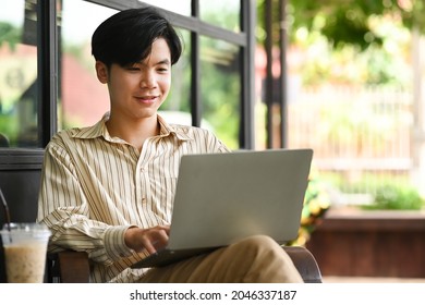 Smiling Young Asian Man Sitting At Outdoor Cafe And Using Computer Laptop.