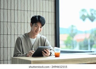 Smiling young Asian man sitting near window at coffee shop and reading book. - Powered by Shutterstock