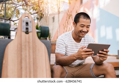 Smiling Young Asian Man Using A Digital Tablet While Sitting Alone Outside On Some Stairs Next To His Long Board 