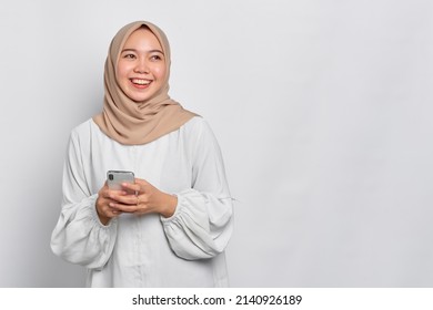 Smiling young Asian Muslim woman using a mobile phone and looking away isolated over white background - Shutterstock ID 2140926189