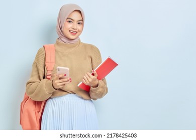 Smiling Young Asian Muslim Woman Student In Brown Sweater With Backpack, Holding Book And Mobile Phone Isolated On White Background. Back To School Concept