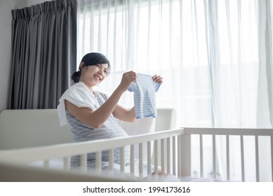 Smiling Young Asian Pregnant Female looking at baby clothes while packing her clothing and getting ready for maternity hospital in bedroom.