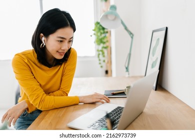 Smiling Young Asian Teen Girl Wearing Headphones Having A Video Call On Laptop. Happy Chinese Pretty Woman Student Looking At Computer Screen Watching Webinar Or Doing Video Chat By Webcam