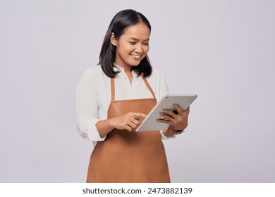 Smiling young Asian woman barista barman employee wearing brown apron working in a coffee shop using digital tablet isolated on white background. Small business startup concept - Powered by Shutterstock