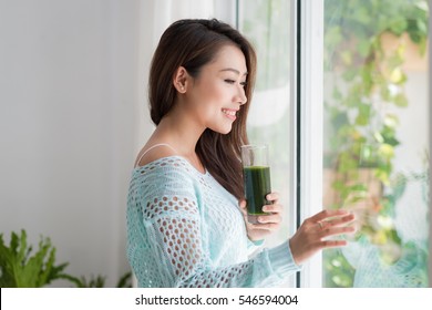 Smiling Young Asian Woman Drinking Green Fresh Vegetable Juice Or Smoothie From Glass At Home